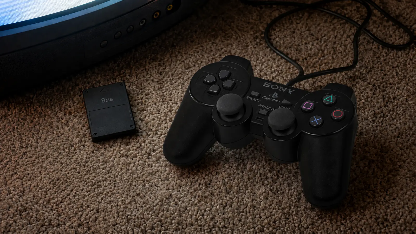 Mando DualShock 2 y tarjeta de memoria de 8 MB sobre una alfombra junto a un televisor CRT.