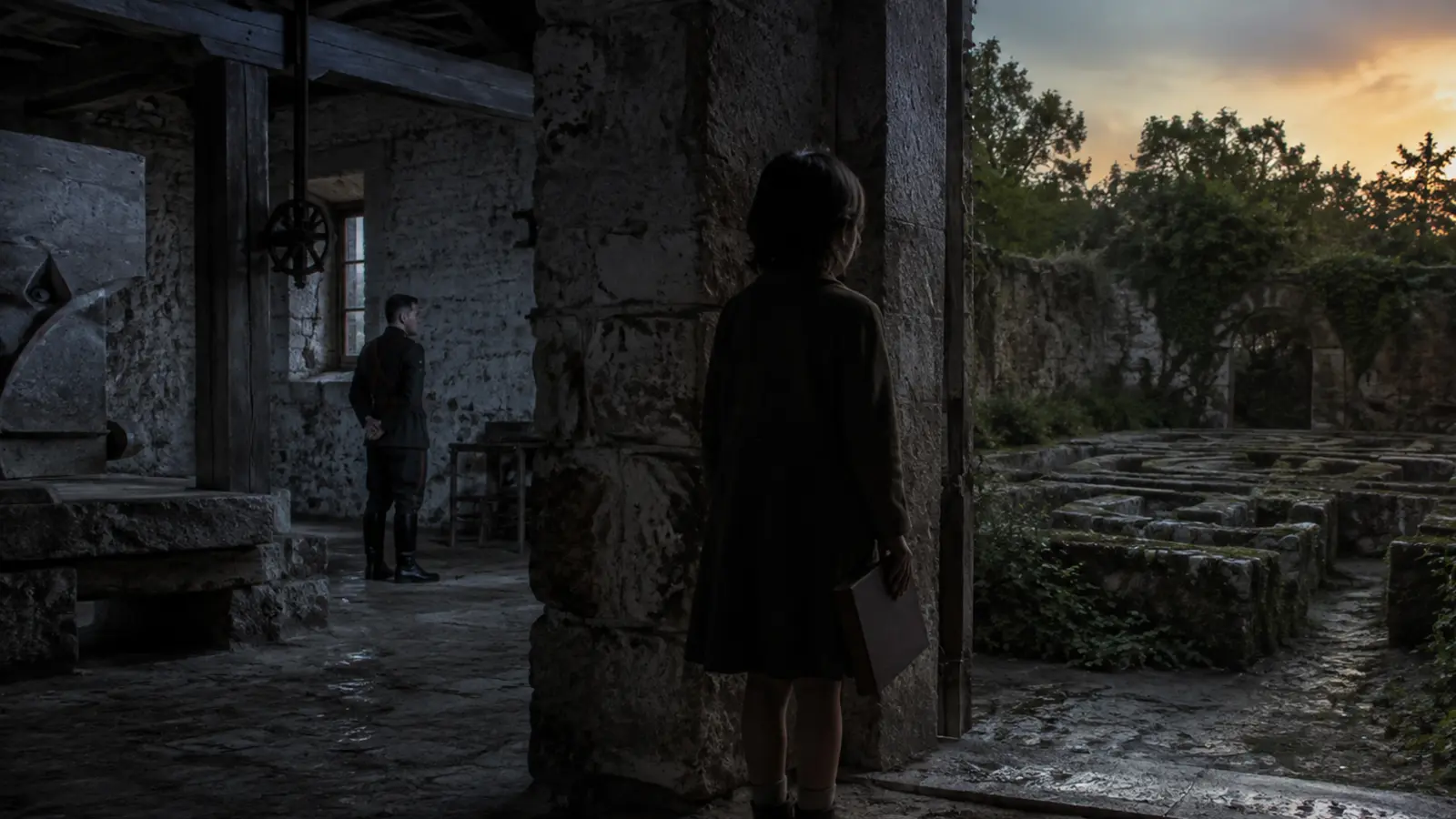 Niña con un libro entre un molino militar y un laberinto de piedra al atardecer.