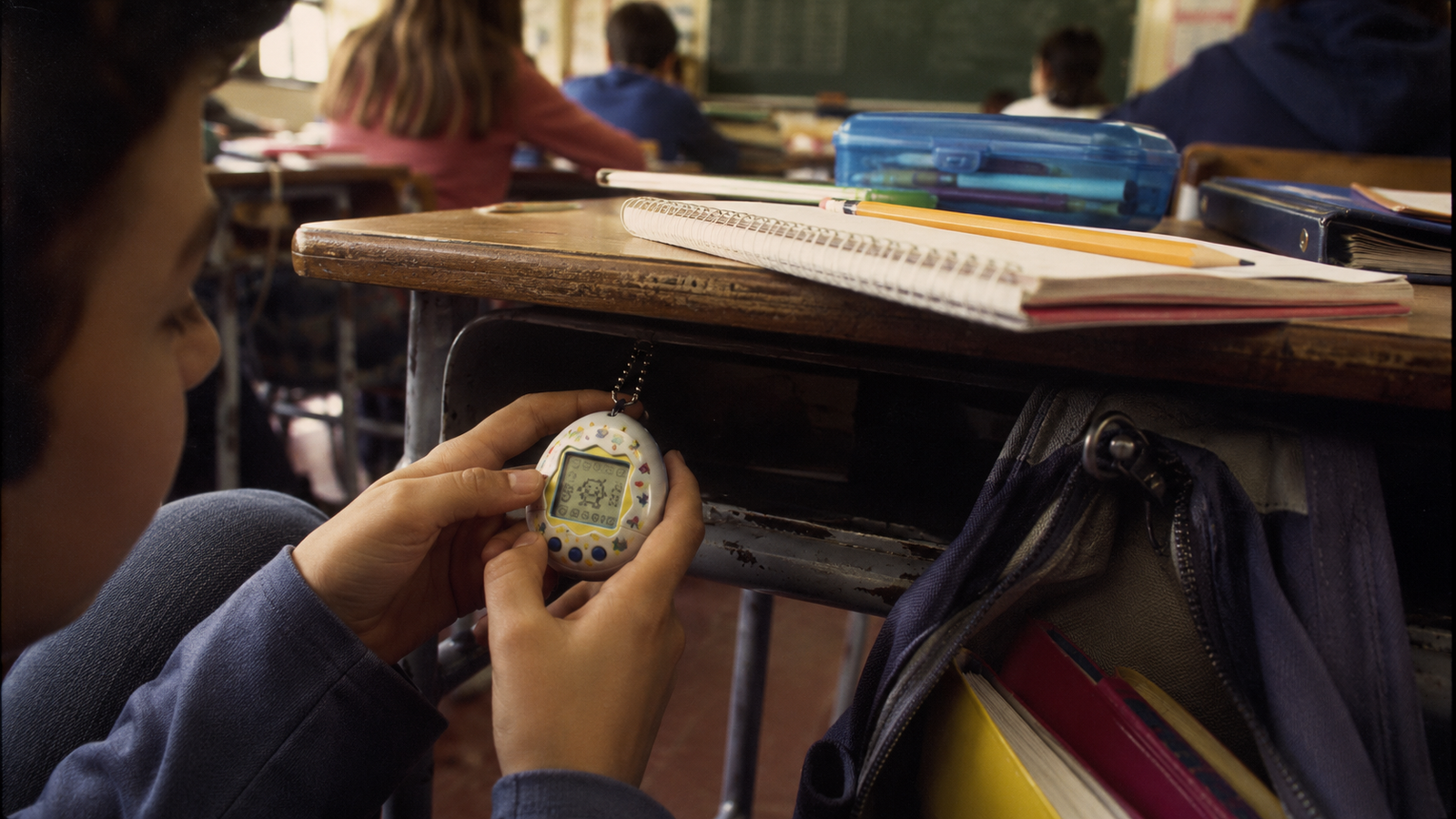 Illustration of a pupil checking a small virtual-pet device below a classroom desk